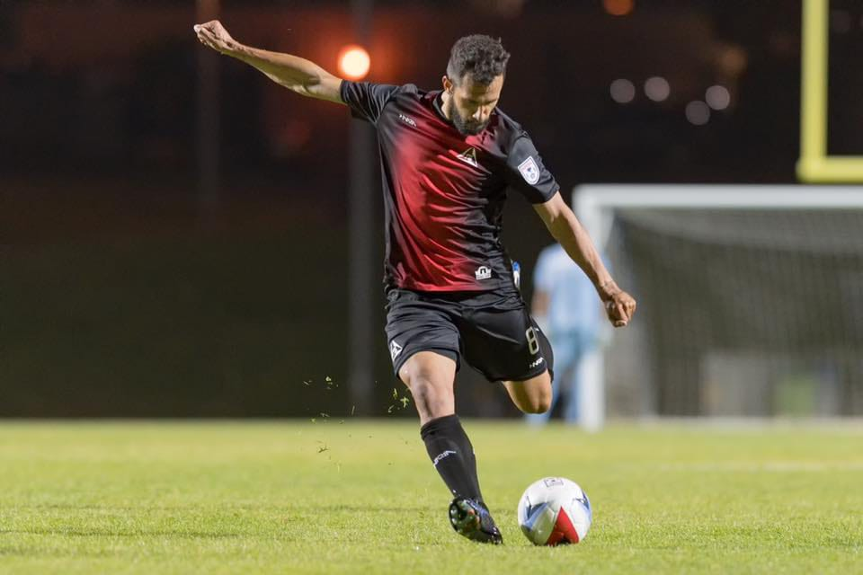 Futbolista con el dorsal 8, vestido de negro y rojo, patea un balón en un campo iluminado de noche. Al fondo se ve una portería y luces desenfocadas.